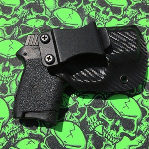 May include: A black handgun with a black carbon fiber holster. The holster has a belt clip and a screw on the side. The gun and holster are on a green background with a skull pattern.