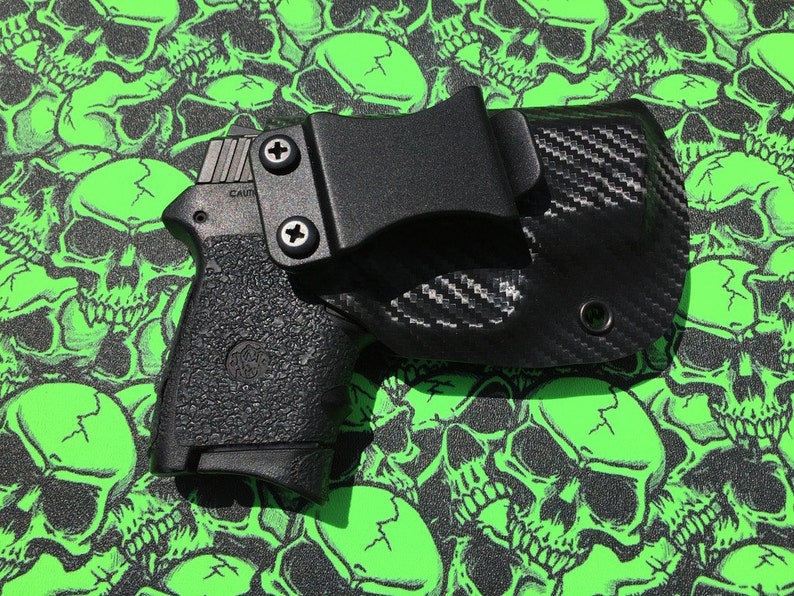 May include: A black handgun with a black carbon fiber holster. The holster has a belt clip and a screw on the side. The gun and holster are on a green background with a skull pattern.