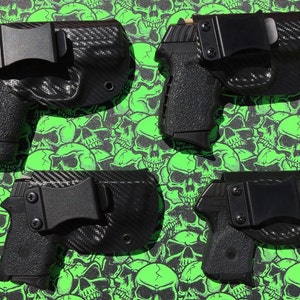 May include: Four black gun holsters with carbon fiber patterns and black clips. The holsters are on a green background with skull designs.