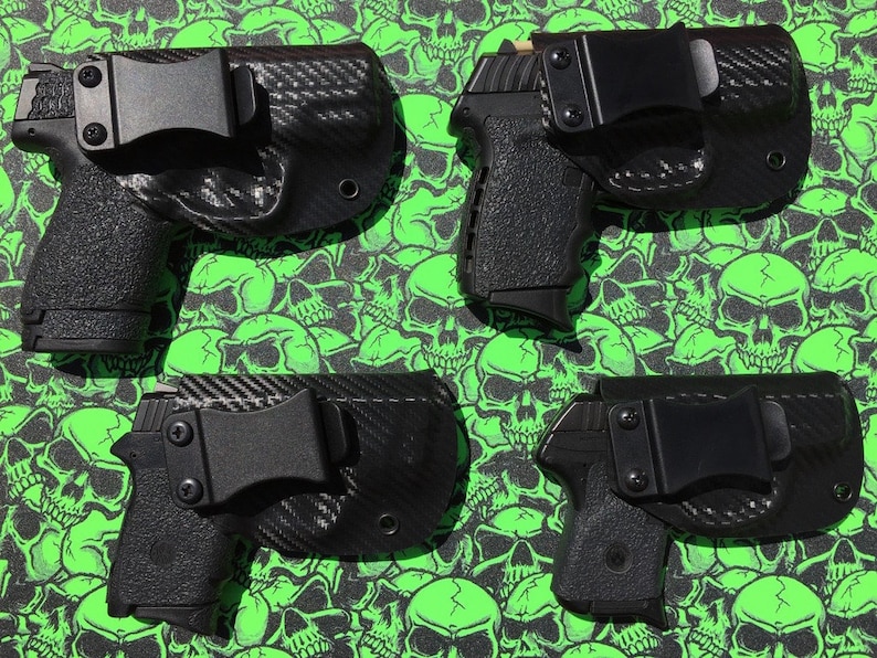 May include: Four black gun holsters with carbon fiber patterns and black clips. The holsters are on a green background with skull designs.