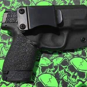 May include: A black handgun with a black holster. The holster has a belt clip and a small hole on the side. The gun and holster are on a green background with a skull pattern.