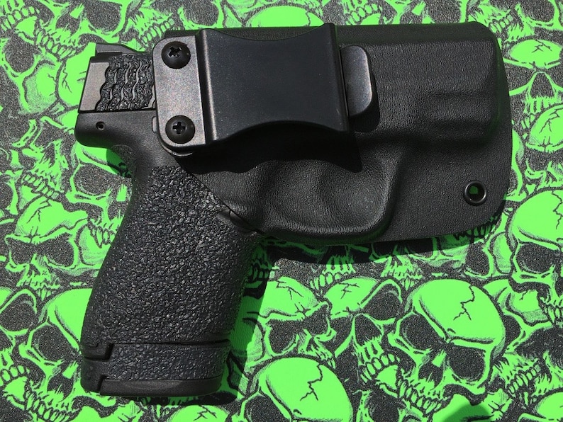 May include: A black handgun with a black holster. The holster has a belt clip and a small hole on the side. The gun and holster are on a green background with a skull pattern.