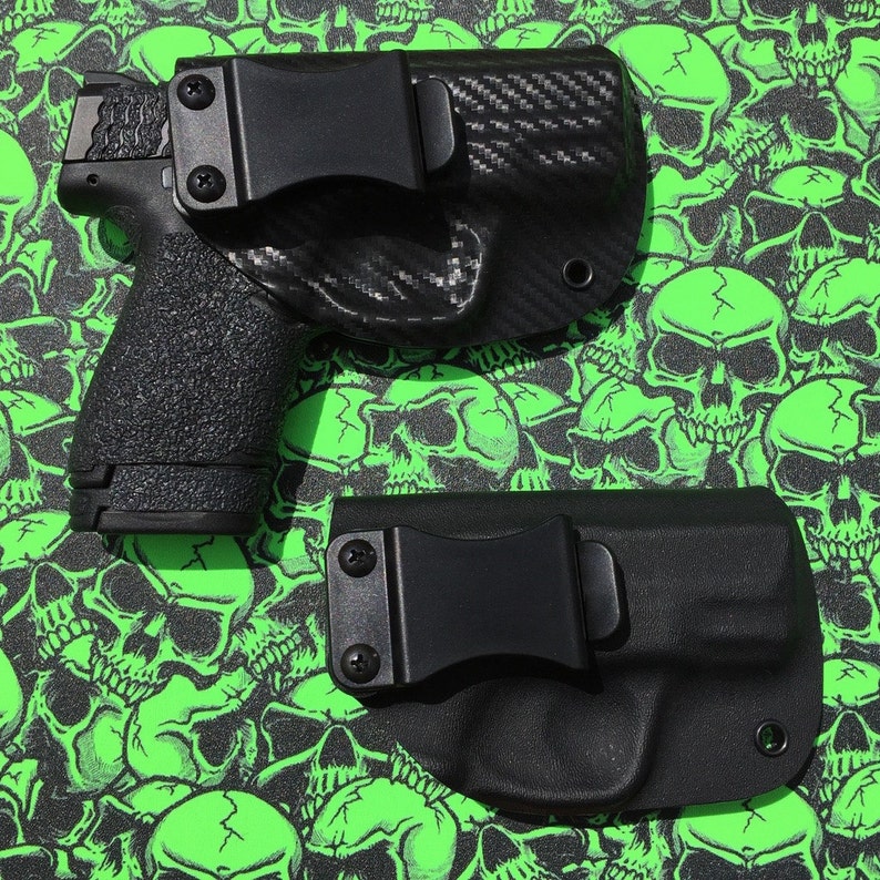 May include: Two black holsters with clips for concealed carry. The holsters are on a green background with a skull pattern.