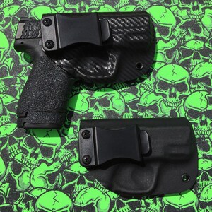 May include: Two black holsters for concealed carry. The holster on the left is made of carbon fiber and has a black clip. The holster on the right is made of a solid black material and has a black clip.