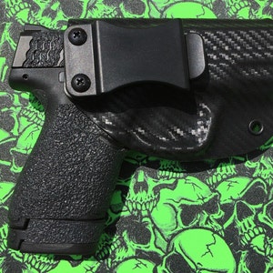 May include: A black handgun with a textured grip is holstered in a black carbon fiber holster with a belt clip.