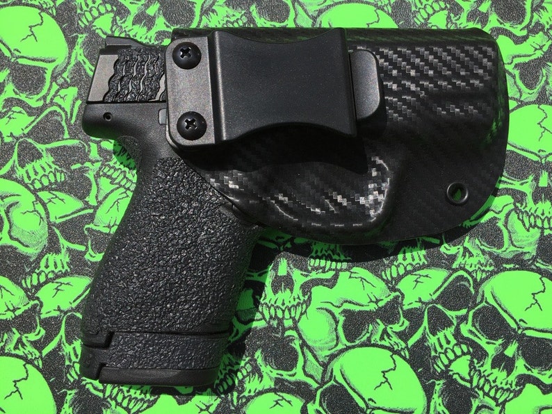 May include: A black handgun with a textured grip is holstered in a black carbon fiber holster with a belt clip.