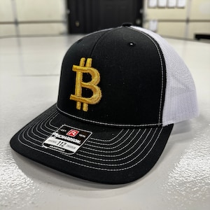 May include: Black and white trucker hat with a gold embroidered Bitcoin symbol. The hat is adjustable and has a Richardson 112 label.