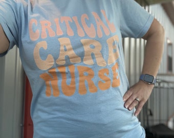 May include: A light blue t-shirt with the text "CRITICAL CARE NURSE" in a retro font with pink, orange, and yellow colors.