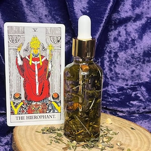 Puede incluir: Una botella de vidrio transparente con una tapa dorada llena de hierbas y flores verdes. Junto a la botella hay una carta del tarot con las palabras "THE HIEROPHANT" y una ilustración de una figura vestida. Los artículos están sobre una rodaja de madera.