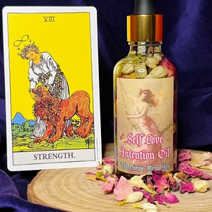 Puede incluir: Una botella de vidrio de Aceite de Intenci&oacute;n de Amor Propio con tapa dorada y gotero. La botella est&aacute; llena de un l&iacute;quido transparente y contiene flores y hierbas secas. La botella est&aacute; sobre una superficie de madera con p&eacute;talos de flores secas esparcidos alrededor. La etiqueta de la botella dice "Self Love Intention Oil" y "The Divine Moon Shop". Una carta de tarot con la imagen de la carta de la Fuerza es visible en el fondo.