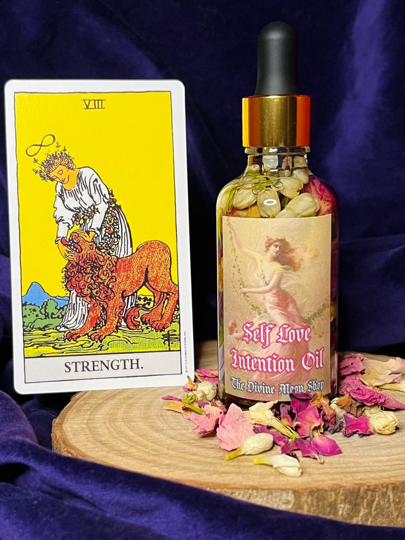 Puede incluir: Una botella de vidrio de Aceite de Intenci&oacute;n de Amor Propio con tapa dorada y gotero. La botella est&aacute; llena de un l&iacute;quido transparente y contiene flores y hierbas secas. La botella est&aacute; sobre una superficie de madera con p&eacute;talos de flores secas esparcidos alrededor. La etiqueta de la botella dice "Self Love Intention Oil" y "The Divine Moon Shop". Una carta de tarot con la imagen de la carta de la Fuerza es visible en el fondo.