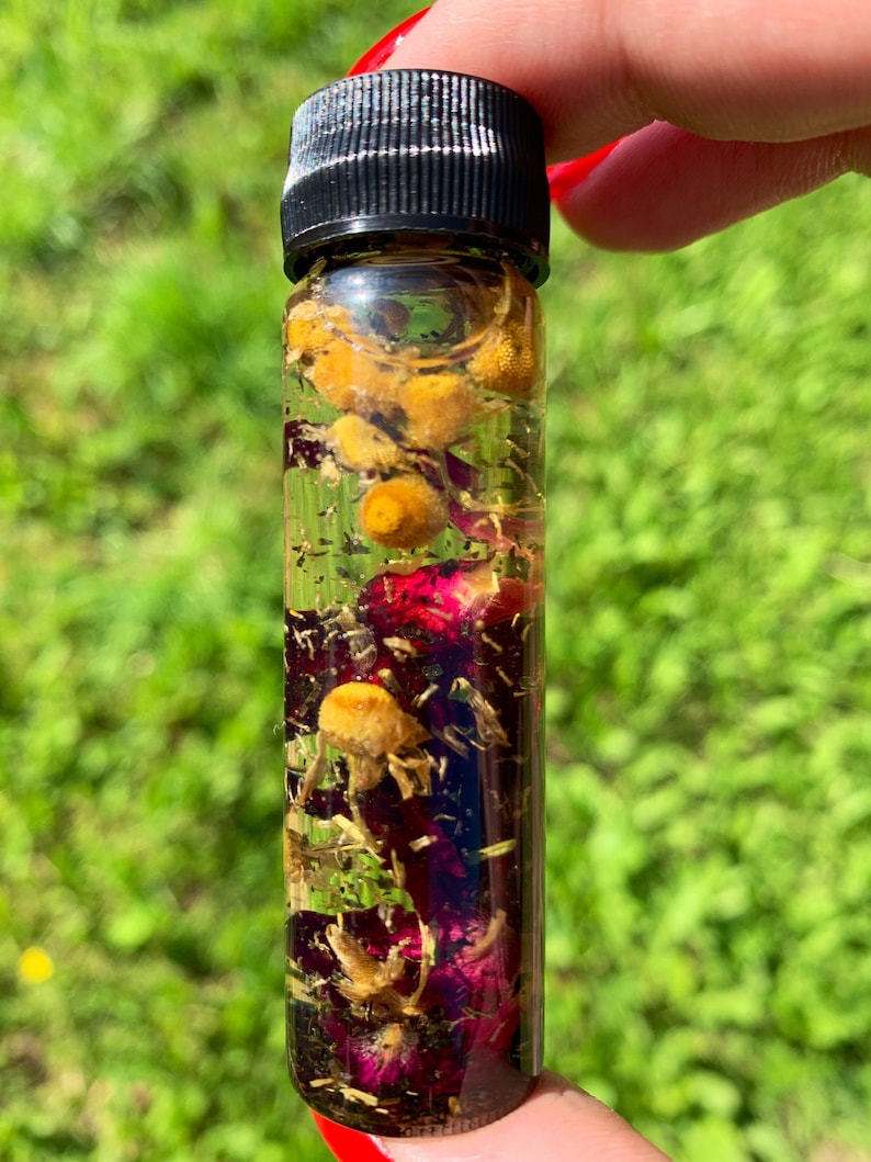 May include: A small glass bottle filled with a clear liquid and dried flowers. The flowers are yellow, pink, and brown. The bottle has a black screw-on cap.