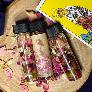 Puede incluir: Tres peque&ntilde;as botellas de vidrio llenas de l&iacute;quido transparente y flores secas. Las botellas tienen tapas negras y etiquetas que dicen "Self Love Intention Oil" y "Divine Moon Spell".