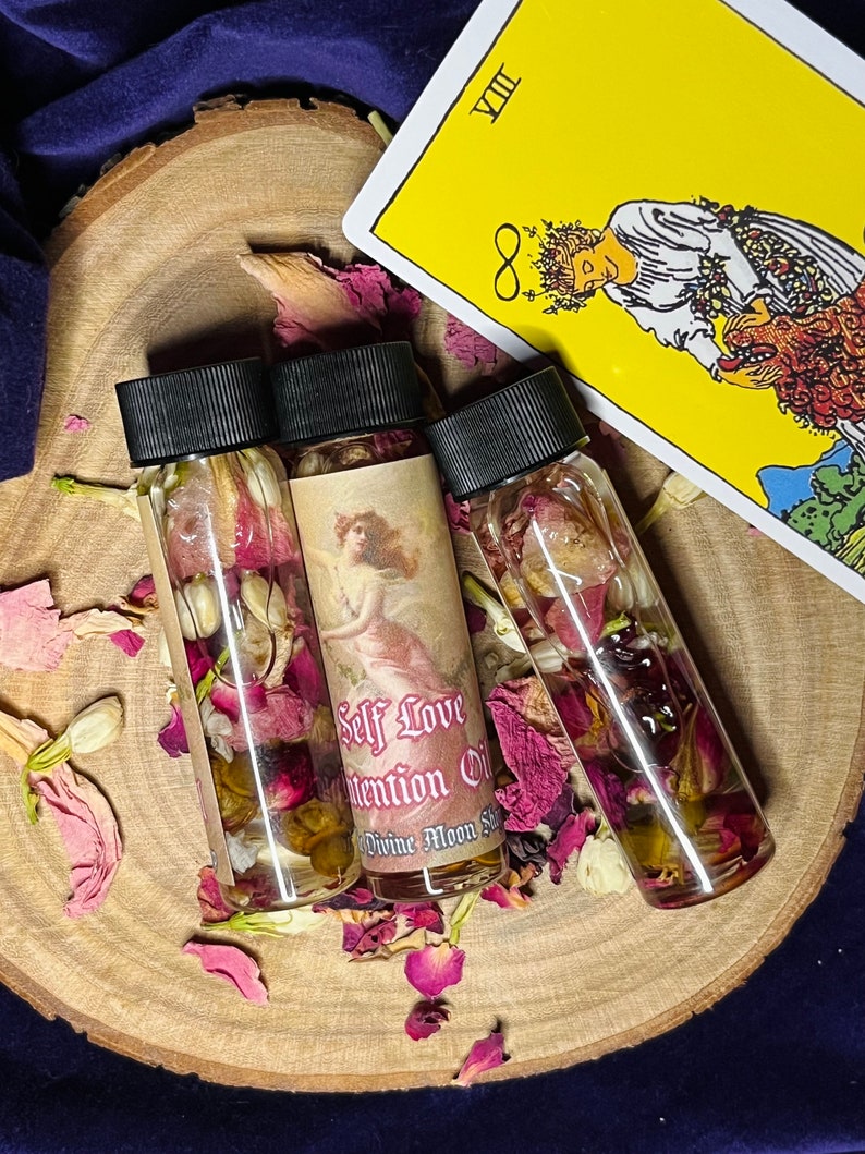 Puede incluir: Tres peque&ntilde;as botellas de vidrio llenas de l&iacute;quido transparente y flores secas. Las botellas tienen tapas negras y etiquetas que dicen "Self Love Intention Oil" y "Divine Moon Spell".