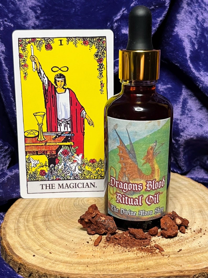 K&ouml;nnte beinhalten: Eine Glasflasche mit Drachenblut-Ritual&ouml;l mit goldenem Deckel und schwarzem Tropfer. Die Flasche hat ein Etikett mit einem roten Drachen und dem Text "Dragons Blood Ritual Oil" und "The Divine Moon Shop". Die Flasche steht auf einer Holzoberfl&auml;che mit einer Tarotkarte, die den Magier zeigt.