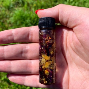 May include: A small glass bottle filled with a clear liquid and dried flowers. The bottle has a black screw-on cap.
