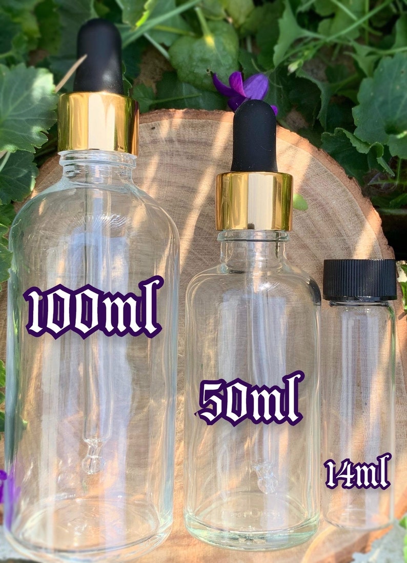 Puede incluir: Tres botellas de vidrio con tapas doradas y goteros. Las botellas son transparentes y tienen etiquetas con el texto "100ml", "50ml" y "14ml" en una fuente g&oacute;tica.