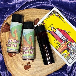 K&ouml;nnte beinhalten: Drei kleine Glasflaschen mit schwarzen Kappen und Etiketten mit der Aufschrift "Dragons Blood Ritual Oil" und "Divine Moon Shop". Die Flaschen stehen auf einer Holzoberfl&auml;che mit einer Tarotkarte im Hintergrund.