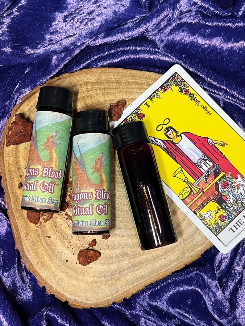 K&ouml;nnte beinhalten: Drei kleine Glasflaschen mit schwarzen Kappen und Etiketten mit der Aufschrift "Dragons Blood Ritual Oil" und "Divine Moon Shop". Die Flaschen stehen auf einer Holzoberfl&auml;che mit einer Tarotkarte im Hintergrund.