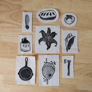 May include: Nine black and white fabric patches with various designs, including a lily, a beehive, a maple leaf, a jug, a skillet, a persimmon, a loaf of bread, a hatchet, and a sprig of lily of the valley.