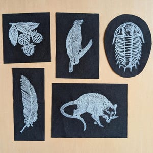 Könnte beinhalten: Fünf schwarze Stoffaufnäher mit weiß gestickten Designs. Die Aufnäher zeigen eine Beerenansammlung, einen Vogel auf einem Ast, ein Trilobitenfossil, eine Feder und ein kleines Tier. Die Aufnäher sind auf einer hellbraunen Oberfläche angeordnet.