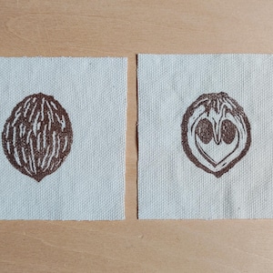 May include: Two white fabric squares with brown printed images of a walnut. The image on the left shows the whole walnut, while the image on the right shows the walnut cut in half.