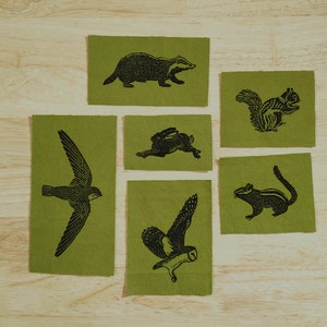 Könnte beinhalten: Sechs grüne Stoffquadrate mit schwarzen Tierillustrationen. Die Tiere sind ein Dachs, ein Eichhörnchen, ein Kaninchen, ein Vogel, ein Mauersegler und ein Streifenhörnchen.
