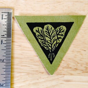 May include: A green fabric triangle patch with a black triangle inside. The black triangle contains a black and white illustration of three leafy plants.