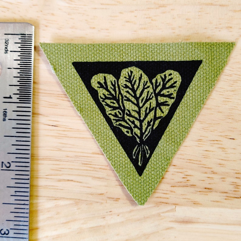 May include: A green fabric triangle patch with a black triangle inside. The black triangle contains a black and white illustration of three leafy plants.