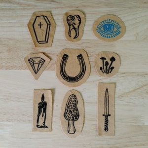 Puede incluir: Nueve parches para planchar con varios diseños, que incluyen un ataúd, un diente, un ojo, una herradura, champiñones, una vela, un champiñón y una daga. Los parches están todos sobre una superficie de madera marrón claro.