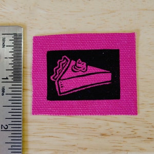 May include: A pink fabric patch with a black square containing a pink and white illustration of a slice of pie.