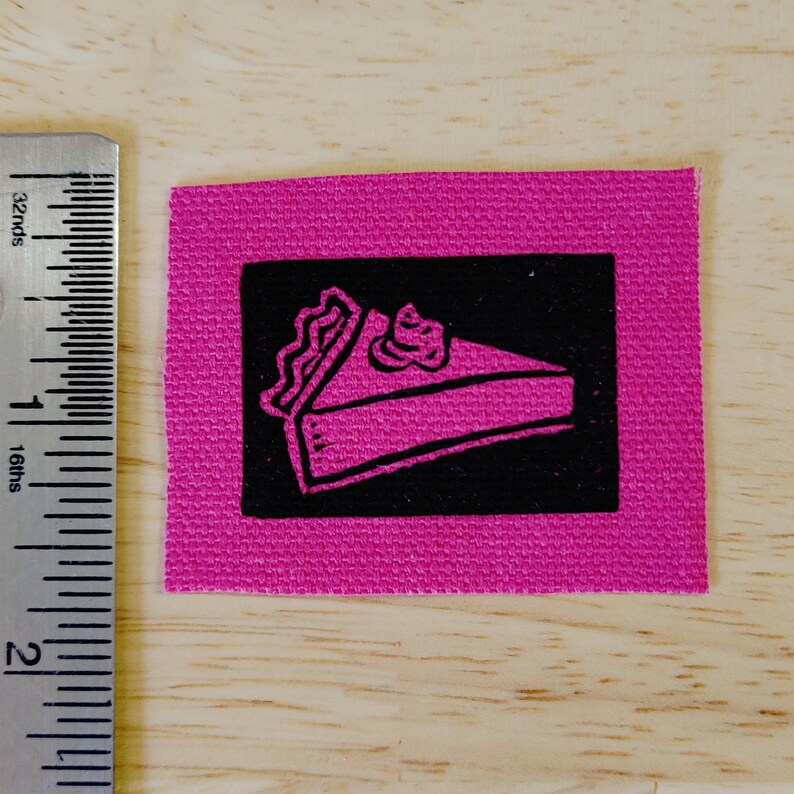 May include: A pink fabric patch with a black square containing a pink and white illustration of a slice of pie.