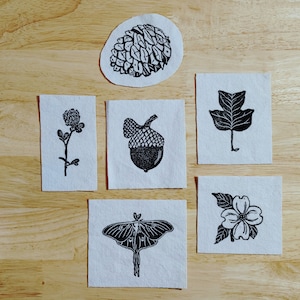 May include: Six black and white fabric patches with nature-themed designs. The designs include a pine cone, an acorn, a leaf, a clover, a moth, and a flower.