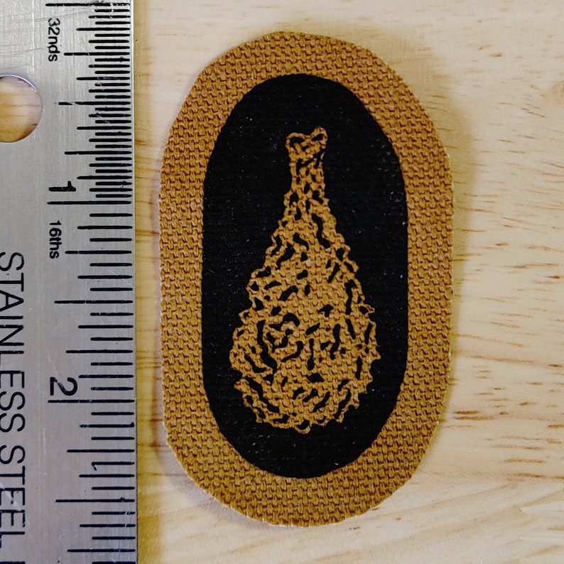 May include: A brown fabric patch with a black oval border. Inside the oval is a black and white image of a pine tree.