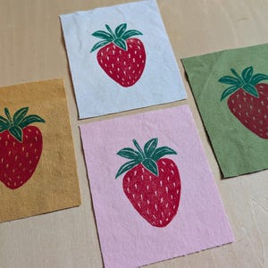 May include: Four fabric squares featuring a red strawberry design with green leaves and stems. The squares are in yellow, white, pink, and green. The strawberries are hand-drawn, creating a rustic aesthetic.