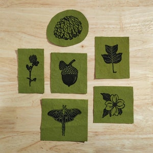 May include: Five green felt patches with black ink illustrations of nature. The patches feature a pine cone, an acorn, a clover, a leaf, and a dogwood flower.