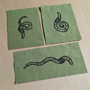 May include: Three olive green fabric patches with black worm illustrations. The patches are rectangular and feature different worm designs, including coiled and straight forms. The illustrations are detailed and appear hand-drawn.