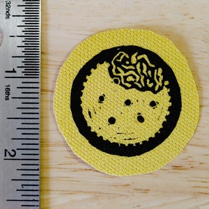 May include: A yellow fabric patch with a black circle design. The circle features a black and white design of a brain with a wavy pattern. There are five black dots around the brain.