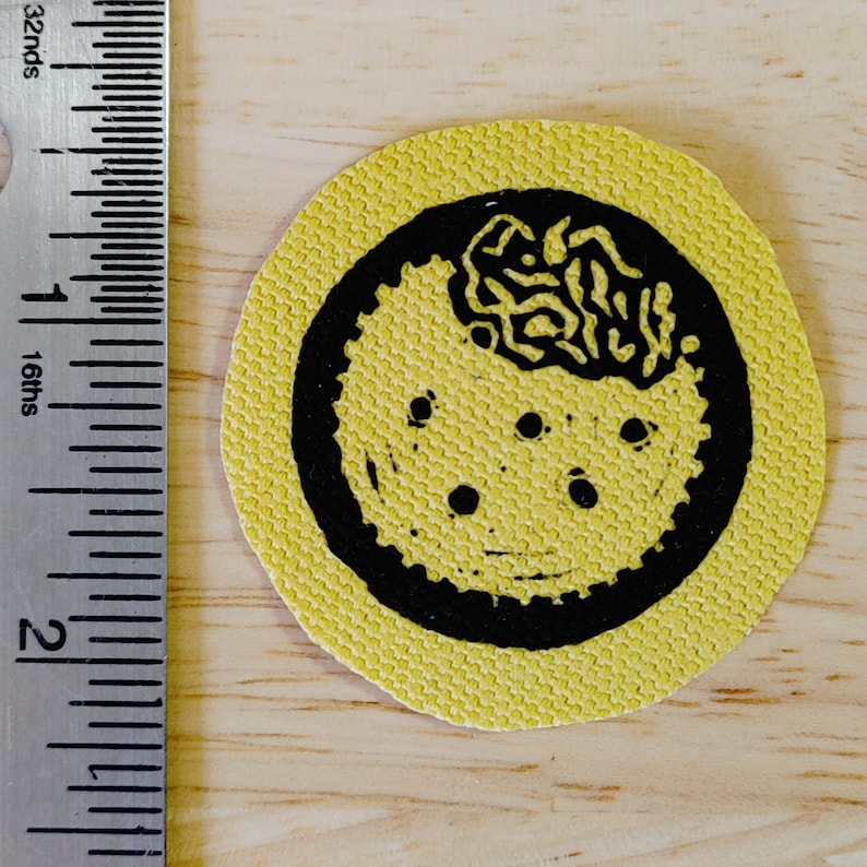 May include: A yellow fabric patch with a black circle design. The circle features a black and white design of a brain with a wavy pattern. There are five black dots around the brain.