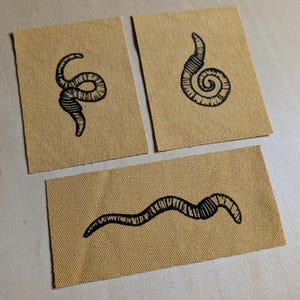 Puede incluir: Tres parches de tela beige con diseños de arte lineal negro. Un parche presenta una forma curva segmentada similar a un gusano. Otro tiene un diseño en espiral, y el tercero tiene un diseño ondulado segmentado. Los parches están dispuestos sobre una superficie clara.