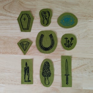 Puede incluir: Un conjunto de parches de tela verde oliva con ilustraciones negras. Los diseños incluyen un ataúd, un diente, un ojo, un diamante, una herradura, setas, una vela, una colmenilla y una daga. Los parches están sobre una superficie de madera clara.