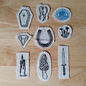 Könnte beinhalten: Zehn Bügelbilder mit verschiedenen Designs, darunter ein Sarg, ein Zahn, ein Auge, Pilze, ein Diamant, ein Hufeisen, eine Kerze, ein Pilz und ein Dolch. Die Patches sind alle weiß mit schwarzer Tinte.