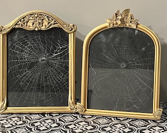 May include: Two ornate gold picture frames with black backgrounds, each featuring a white spiderweb design. One frame has a decorative top, the other has a butterfly motif. The frames are displayed on a black and white patterned surface.
