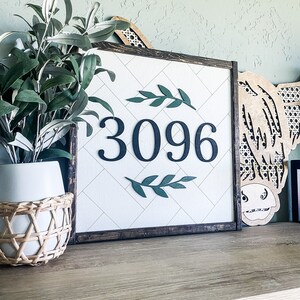 May include: A framed house number sign with the number 3096 in dark blue. The sign has a white background with a geometric pattern and is framed in dark brown. Green leaf accents are above and below the numbers. A decorative piece and a plant are in the background.