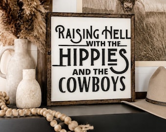 May include: A square wooden sign with a dark brown frame and a white background featuring raised black lettering that reads Raising Hell with the Hippies and the Cowboys. The sign has a clean, bold font style with distinct, layered text elements.