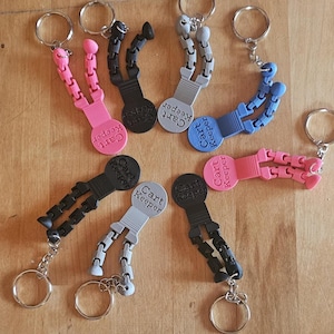 Puede incluir: Una colección de llaveros "Cart Keeper" coloridos. Los llaveros presentan un diseño de brazo segmentado en rosa, negro, gris y azul, unido a una base circular con el texto "Cart Keeper". Cada llavero tiene un llavero plateado.