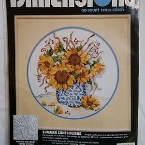 May include: A Dimensions no count cross stitch kit titled Summer Sunflowers featuring a printed design of yellow sunflowers in a blue and white spongeware vase. The package indicates a 17 inch by 17 inch frame size and a 14 inch diameter mat.