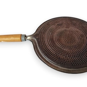 May include: A round, dark brown metal heat diffuser plate featuring a dense pattern of small, uniform perforations across its surface. The circular plate is attached to a light-colored wooden handle with a grey collar and a thin leather hanging cord.