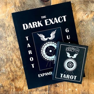 May include: A black guidebook titled "The Dark Exact Tarot: Expand Your Understanding" with a black and white illustration of a compass and wings. A smaller black box with the same title and a similar compass illustration is in front of the guidebook.