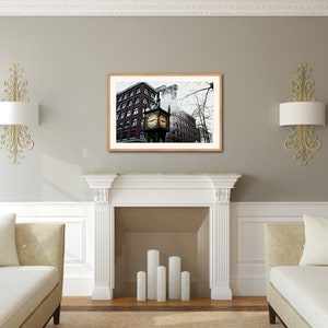 May include: A framed photograph of a clock tower in a city setting. The clock tower is brick with a gold clock face and is surrounded by other buildings. The photo is in a brown frame and is hanging on a gray wall.